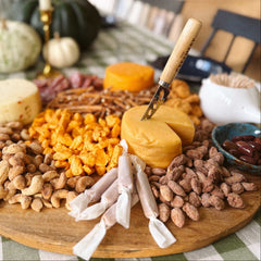 Assorted cheeses and cheese curds from The Creamery, candied nuts, caramels, pretzels and olives on a wooden board with The Creamery Wooden Cheese Knife, set on a table 