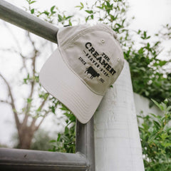The Creamery Stone Cool Fit Hat hanging from a fence outside